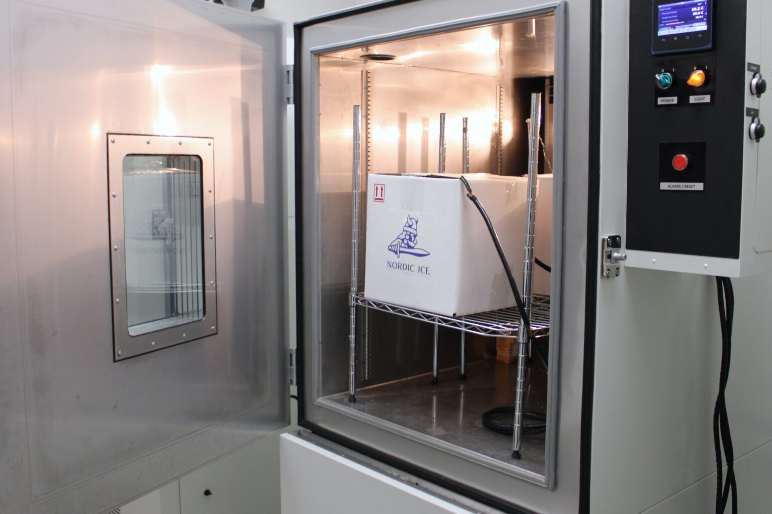 An industrial refrigeration chamber with the door open, showing a white cardboard package on a metal grid. The package reads 'Nordic Ice'. Next to the chamber is a control panel with buttons and a digital display, indicating that it is automated equipment.